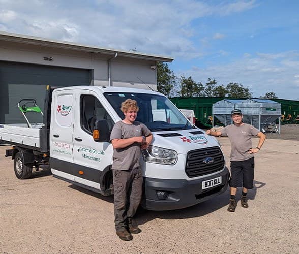 Two Avery grounds maintenance operatives beside a branded tipper truck loaded with mowers