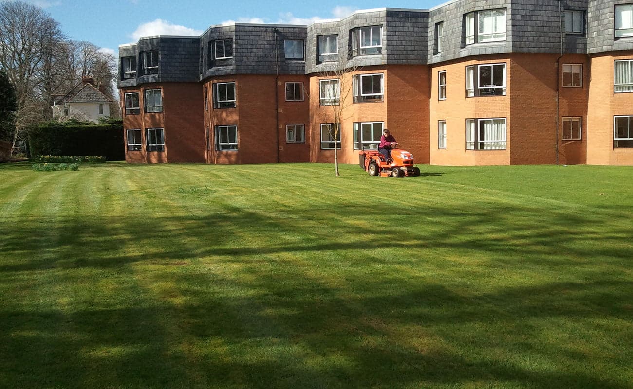 Ride-on mower cutting a large striped lawn in front of a later-living apartment block